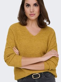 Senfgelber, gerippter Strickpullover mit V-Ausschnitt, entspannter Passform und langen Ärmeln. Kombiniert mit einem schwarzen Gürtel mit goldener Schnalle.