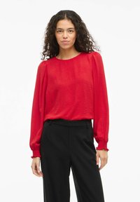 Blusa roja con textura, de mangas largas y abullonadas, con puños fruncidos, combinada con pantalones negros de cintura alta. Cuello redondo simplista.