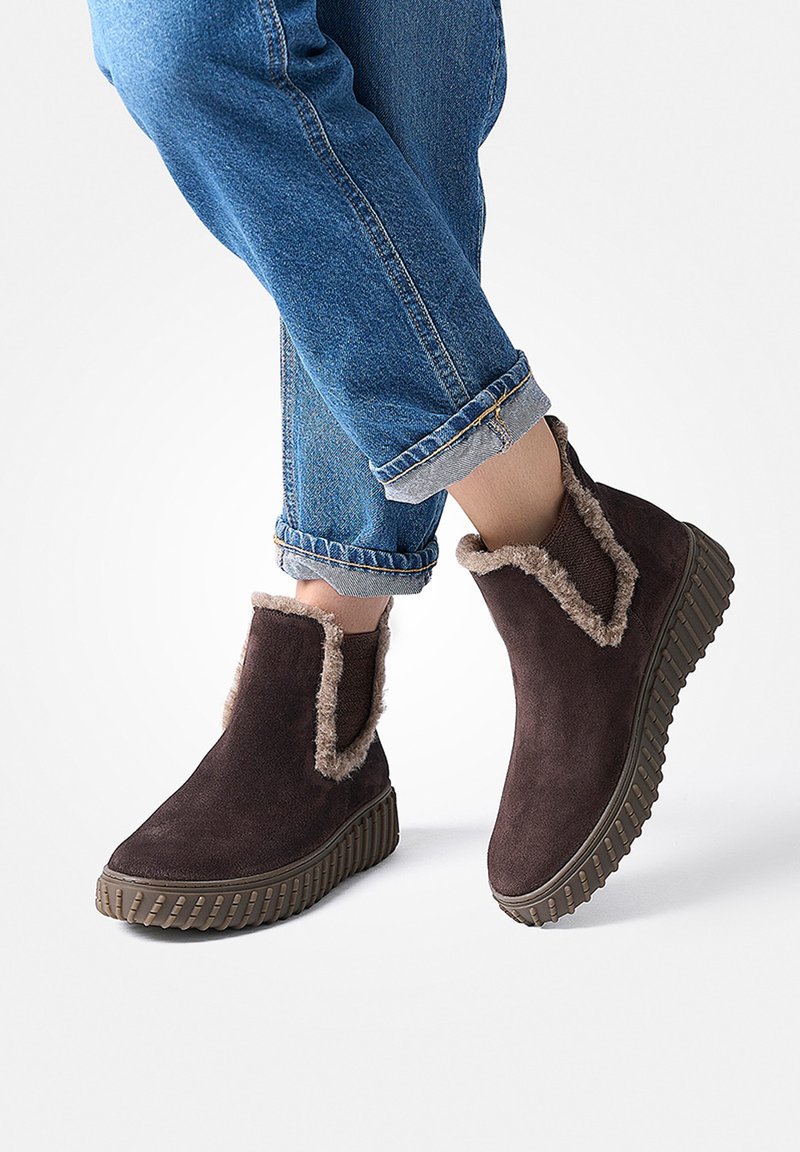 Braune Wildleder-Stiefeletten mit einer strukturierten Gummisohle, die mit einem weichen Fleecebesatz und seitlichen Ausschnitten versehen sind. Getragen mit hochgekrempelten blauen Jeans.