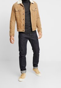 Veste en velours côtelé beige avec un col en peau lainée blanche, pull noir, jean foncé et baskets montantes beiges, présentant une tenue décontractée en couches.