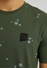 Zelené tričko s krátkými rukávy vyrobené z bavlny, s světle modrými kruhovými vzory a černou obdélníkovou nášivkou s šikmým designem.