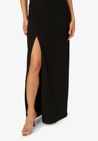 Černé maxi šaty s rozparkem až na stehna, vyrobené z hladkého materiálu. Transparentní sandály na vysokém podpatku s tenkými pásky doplňují celek.