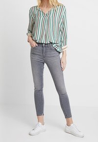 Blusa a righe verde e bianca con scollo a V e maniche piegate, abbinata a jeans skinny grigi e sneakers bianche.