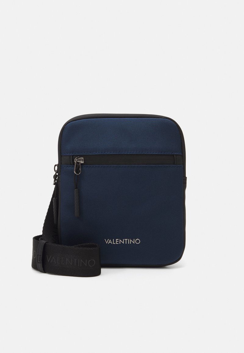 Sac bandoulière marine en tissu texturé, avec bord noir, poche zippée à l'avant et sangle ajustable arborant un logo en relief. Design compact et rectangulaire.