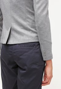 Gros plan d'une personne portant un blazer gris ajusté avec des poignets boutonnés et un pantalon bleu marine foncé, vu de l'arrière de la taille jusqu'au milieu de la cuisse.