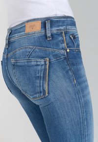 Jean en denim dans un délavage bleu clair avec une taille mi-haute, des coutures dorées et une patch en cuir distinctif à l'arrière.