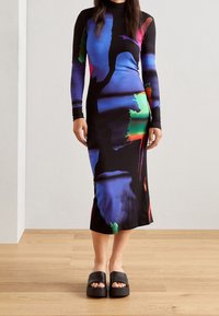 Femme portant une robe noire à manches longues et col montant avec des motifs abstraits bleus, verts et rouges, assortie à des sandales plateformes noires ouvertes.