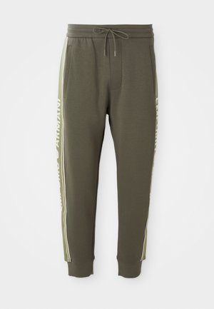 Pantalons de survêtement vert olive en tissu doux avec une taille à cordon de serrage, présentant des rayures sur les côtés et des lettres blanches le long des coutures extérieures.