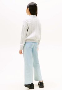 Hellgrauer Strickpullover mit Kabelmuster und geripptem Kragen, kombiniert mit hellblauen Schlagjeans und schwarzen klobigen Schuhen. Sichtbare Designdetails auf der Rückseite.