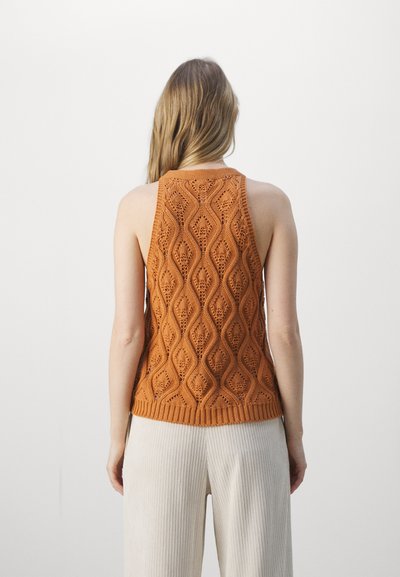 Virkat, ärmlös orange topp med ett vågmönster och ribbad nederkant. Modellen bär krämfärgade, plisserade vida byxor.