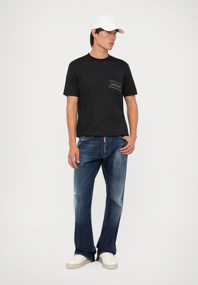 T-shirt preta de mangas curtas com um bolso frontal e riscas brancas, combinada com jeans de perna direita azul desbotados e ténis brancos.