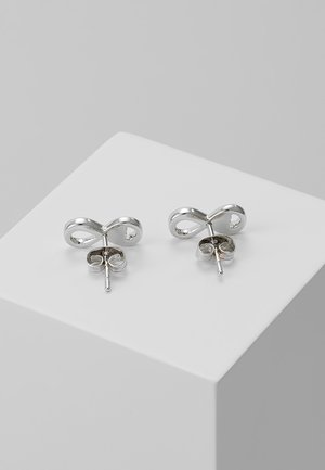 Silberne Ohrstecker in Form von Unendlichkeitssymbolen, die auf einem weißen Sockel vor einem grauen Hintergrund präsentiert werden.