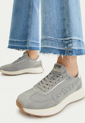 Persona che indossa jeans azzurri con orlo sfrangiato e scarpe da ginnastica grigie G-Star in mesh con suole bianche, in piedi su una superficie bianca.
