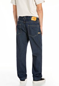 Mørkeblå denimjeans med en afslappet pasform, der har to baglommer, orange syninger og et gult mærke på livbeklædningen.