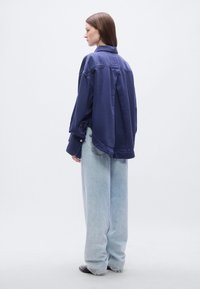 Ναυτικό υπερμεγέθες πουκάμισο από τζιν με γιακά πουκαμίσου, μακρυά μανίκια και διακοσμητικά κουμπιά, σε συνδυασμό με ανοιχτό μπλε τζιν παντελόνι με φαρδύ πόδι και μαύρα παπούτσια.