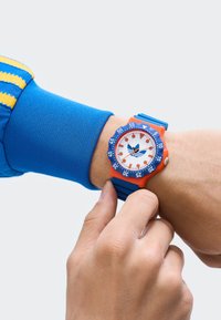 Erinev värviline kell valge näo, oranži korpuse, sinise raami ja rihmaga. Omab julgeid tunni markereid ja emblemid keskel.