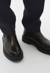 Pruunist nahast Chelsea saapad, millel on sile viimistlus, ümar varvas ja must kummist tald, on kombineeritud tekstureeritud tumesiniste pükstega.