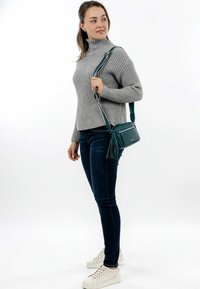 Teal crossbody bag with a textured finish, featuring a long adjustable strap and a tassel accent, paired with a gray ribbed sweater and jeans.