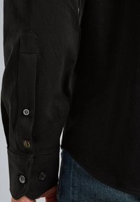 Czarna koszula z długim rękawem z zapięciem na guzik, z eleganckim wykończeniem i subtelną metką logo na rękawie. Materiał wydaje się gładki i trwały.