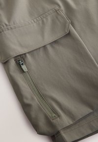 Gros plan sur un tissu kaki avec une poche zippée sur le côté et une poche à rabat, présentant une texture lisse, des coutures renforcées et des accents vert olive.