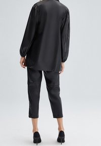 Kvinna i svart satinblus med långa ärmar och korta byxor, matchat med svarta högklackade skor, står framför en neutral bakgrund.