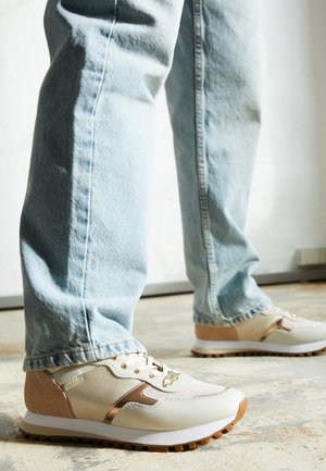 Person iført lyseblå jeans og beige sneakers står på en lysefarvet betongulv.
