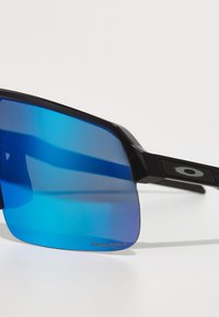 Gafas de sol negras con forma rectangular, lentes con degradado azul y acentos laterales texturizados. Presenta la marca "PRIZM" en la lente.