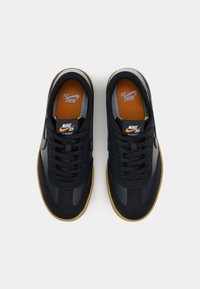 Sapatilhas pretas em couro com uma textura lisa, apresentando uma sola de borracha natural, atacadores pretos e uma palmilha laranja com o logótipo "Nike SB".