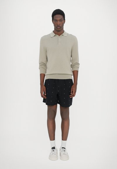 Beige langärmliges Polohemd aus Strickstoff, kombiniert mit schwarzen Shorts, die kleine, hellfarbige geometrische Muster aufweisen, und weißen Sneakern.