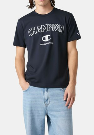 Giovane uomo che indossa una T-shirt Champion blu navy con logo e scritta bianchi, abbinata a jeans azzurri, con le mani parzialmente nelle tasche.