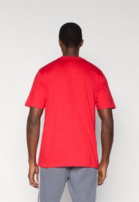 Κόκκινο κοντομάνικο t-shirt με στρογγυλή λαιμόκοψη, κατασκευασμένο από λείο ύφασμα. Η πίσω πλευρά είναι απλή χωρίς ορατά λογότυπα ή σχέδια, φορεμένο με γκρι παντελόνι.