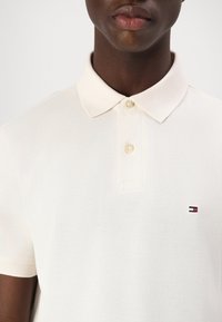 Gros plan d’une personne portant un polo blanc à manches courtes avec une patte à deux boutons et un petit logo Tommy Hilfiger sur la poitrine.