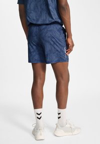 Marinblåa träningsshorts med subtil ruggad textur, åtsittande design och logotypdetalj på sidan, bärs tillsammans med vita sneakers och mönstrade strumpor.