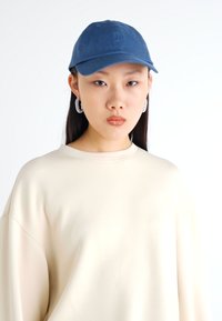 Blue cotton cap with a curved brim, worn with a cream oversized sweatshirt. Model features silver hoop earrings. Smooth fabric texture.