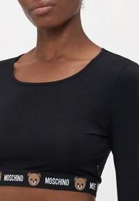 Nærbilde av en person som har på seg en svart crop top med et Moschino-merket elastisk bånd som har teddy-bjørnansikter langs kanten.