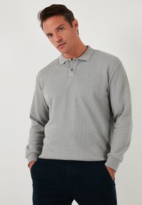 Polo de culoare gri deschis cu mânecă lungă, cu textură canelată, având guler și placket cu două butoane, asociat cu pantaloni închiși la culoare.
