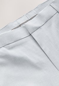 Ceinture de pantalon en tissu gris clair avec une fermeture invisible à crochet et barre et un détail de surpiqûre visible.