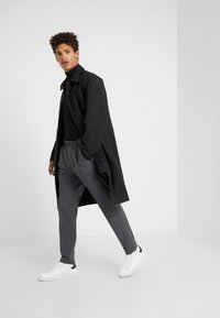 Schwarzer langer Mantel, tailliertes schwarzes Rollkragenoberteil, graue Hose und weiße Sneakers. Modell in Bewegung vor einem schlichten weißen Hintergrund.