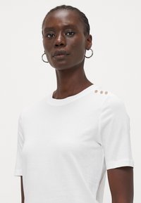 White cotton t-shirt with short sleeves, featuring three decorative buttons on the left shoulder. Smooth texture and classic crew neck design.