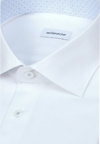 Gros plan sur le col d'une chemise blanche avec deux boutons fermés et une étiquette de marque Seidensticker visible à l'intérieur du col.