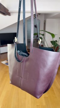 Leren totebag in diep bordeauxrood met een gladde textuur, twee zwarte hengsels en goudkleurige details. Heeft een minimalistisch ontwerp en een open bovenkant.