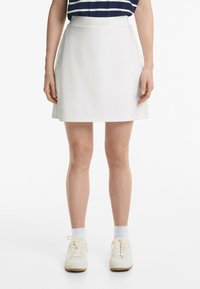 Jupe A-line blanche avec une finition lisse, longueur mi-thigh et poches latérales, associée à des baskets blanches et des chaussettes de cheville côtelées.