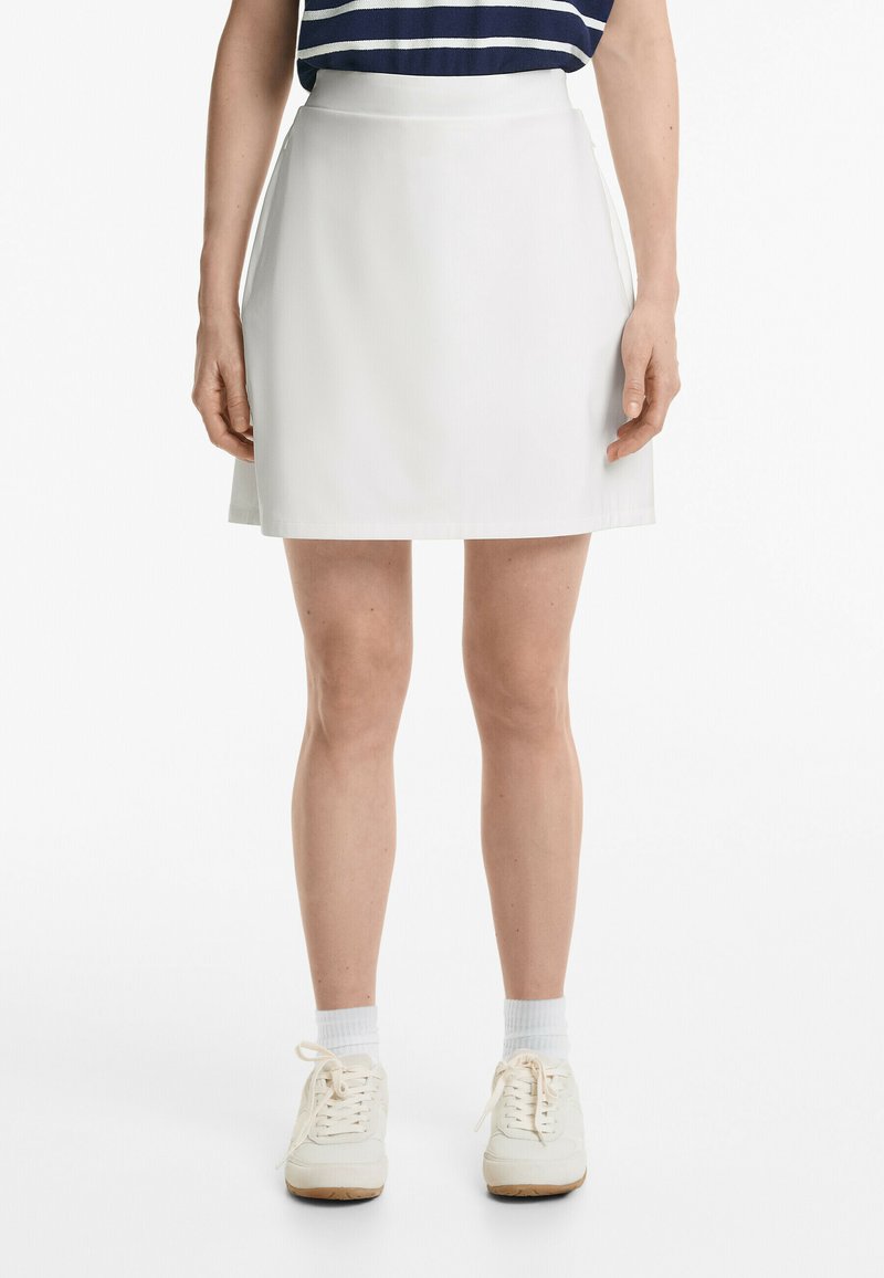 Jupe A-line blanche avec une finition lisse, longueur mi-thigh et poches latérales, associée à des baskets blanches et des chaussettes de cheville côtelées.