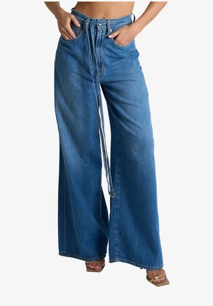 Jeans in denim blu a gamba larga con vita alta, passanti per cintura, tasche anteriori; indossati con sandali con punta quadrata e vista parziale del torso con le mani nelle tasche.