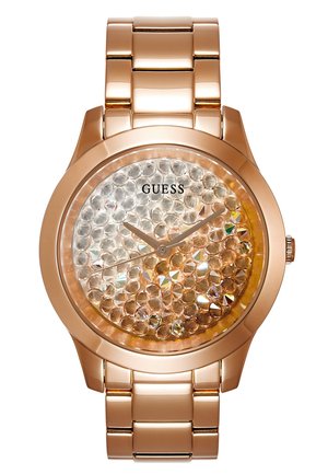 Zegarek z różowego złota z tarczą ozdobioną kryształkami, bez cyfr, z cienkimi wskazówkami i logo GUESS pod pozycją godziny 12.