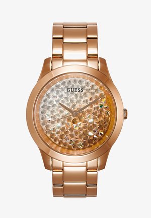 Montre en métal rose doré avec cadran orné de cristaux clairs, sans chiffres, aiguilles fines, et logo GUESS sous la position 12 heures.