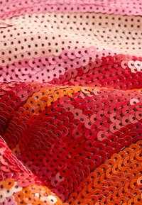 Primer plano de una tela densamente cubierta de lentejuelas brillantes y superpuestas en tonos de rosa, rojo y naranja que crean un patrón texturizado.