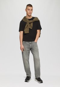 T-shirt noir à manches courtes, sweat à capuche vert olive noué autour des épaules, jeans gris clair à jambe droite et baskets de sport noires.
