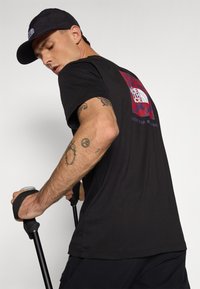 Tricou negru cu mânecă scurtă, având logo-ul The North Face în roșu și albastru pe spate, asortat cu pantaloni scurți negri și ținând bețe de trekking.