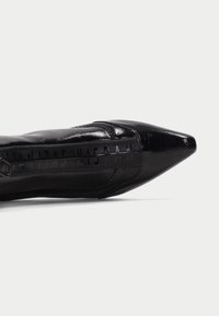 Scarpa nera con punta affusolata realizzata in materiale pelle verniciata lucida, con zip laterale e dettagli cuciti. Suola piatta e design elegante.